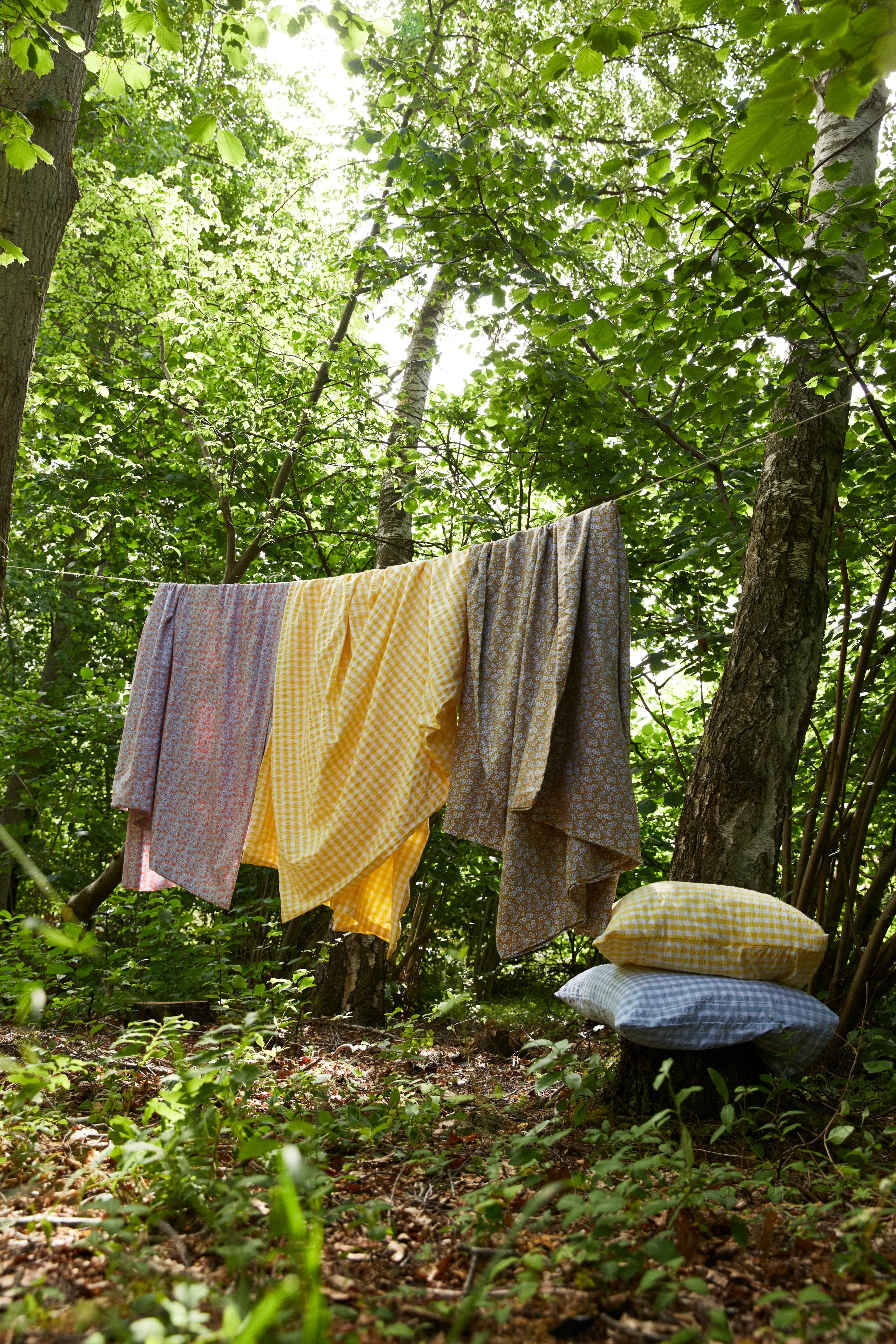 JUNA tekstiler henger til tørk mellom trær i en solrik skog – fargerike mønstre i naturen.