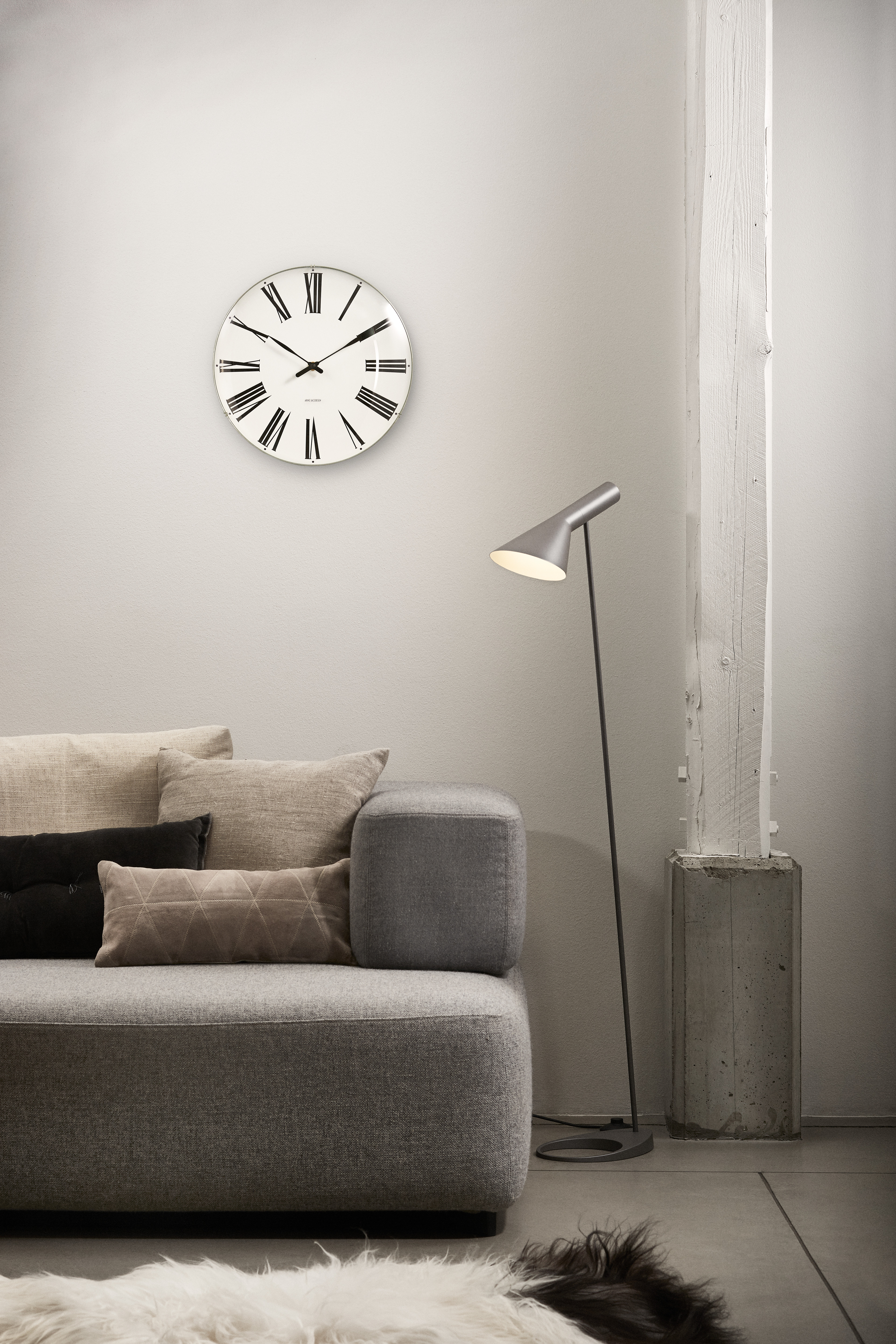 An Arne Jacobsen roman clock hanging abov a grey sofa with beige pillows.