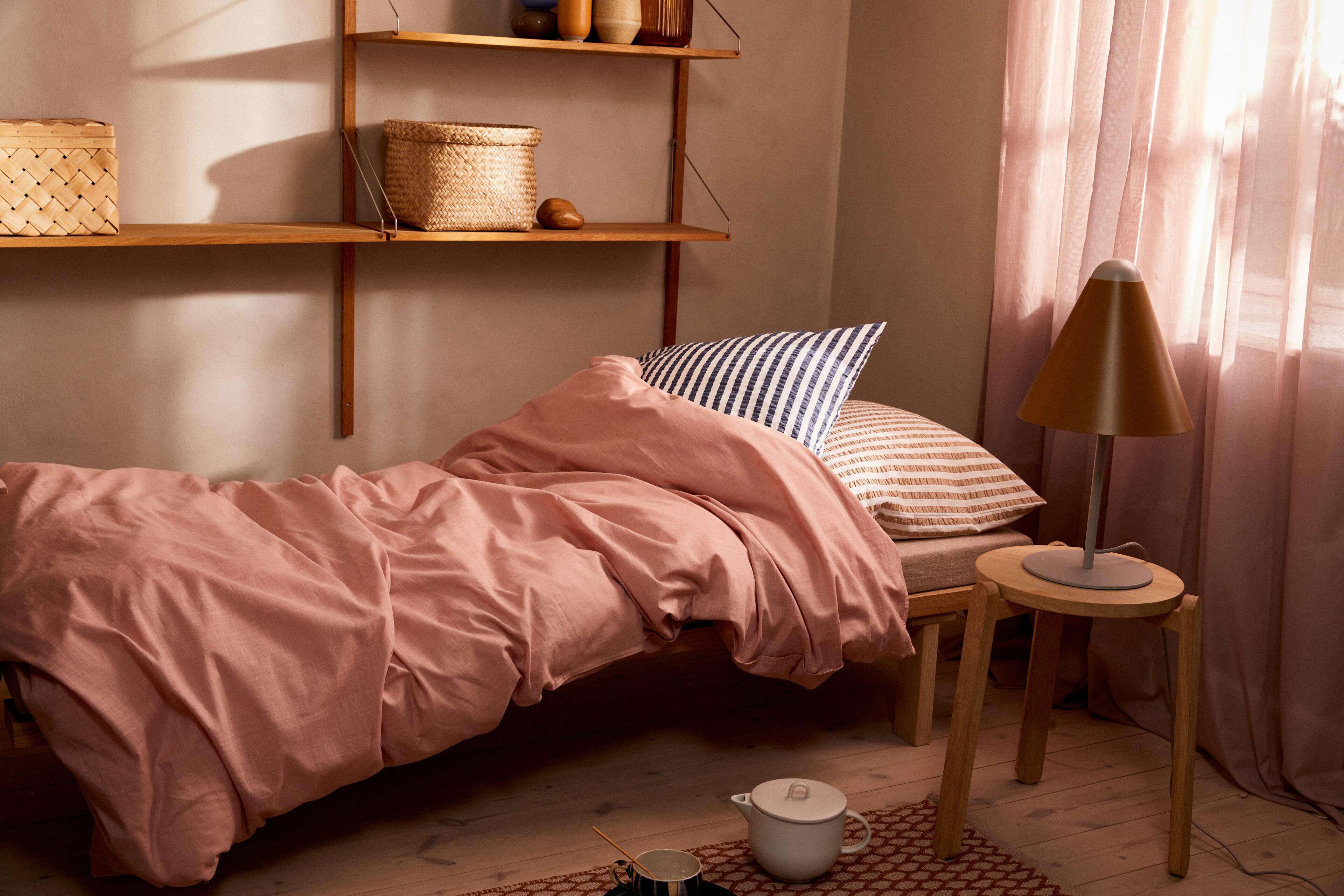 Bett mit Bettwäsche von JUNA im Zimmer mit Hocker, Lampe und rosa Vorhängen