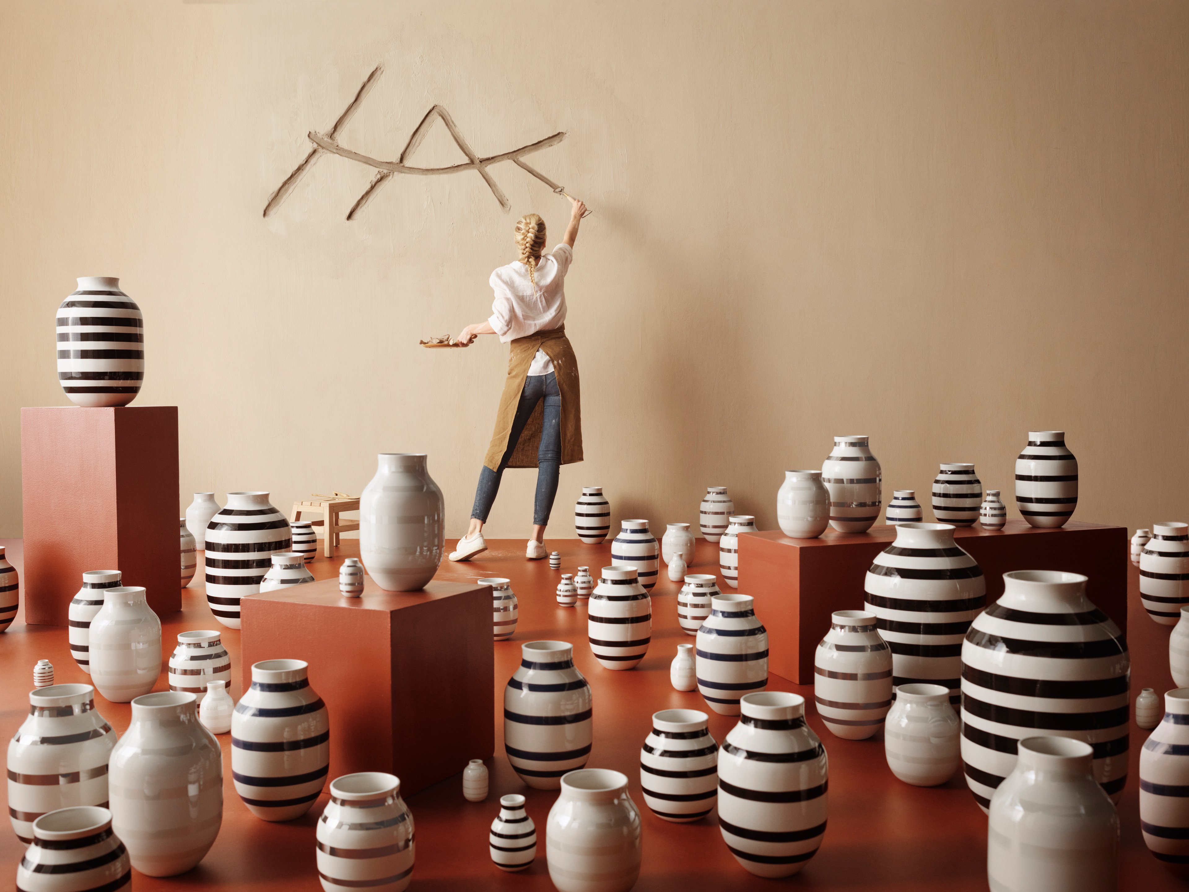 Woman paints Kähler's HAK logo on wall among Omaggio vases