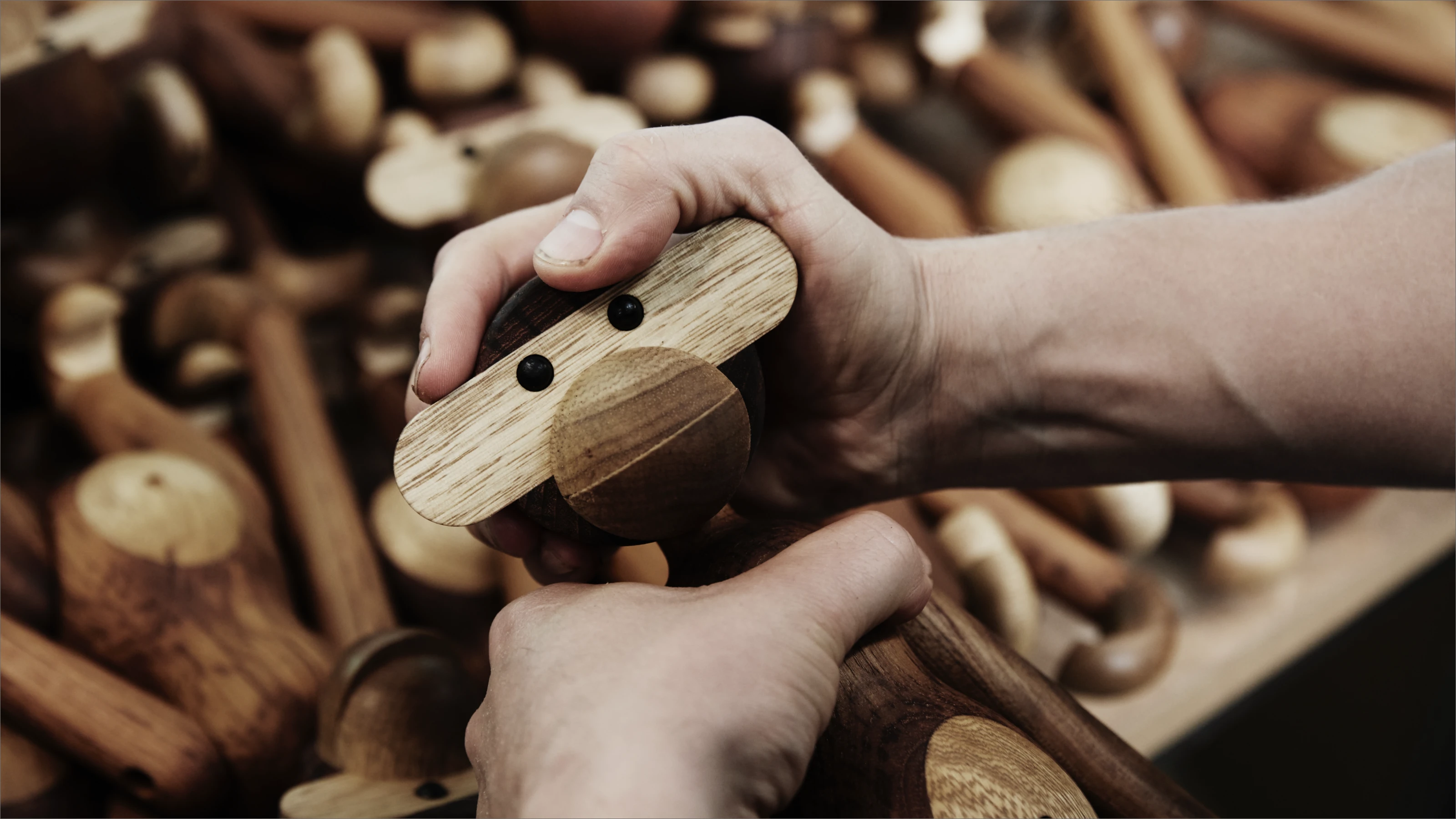 Kay Bojesen Monkey is held in two hands, with a stack of wooden monkeys in the background