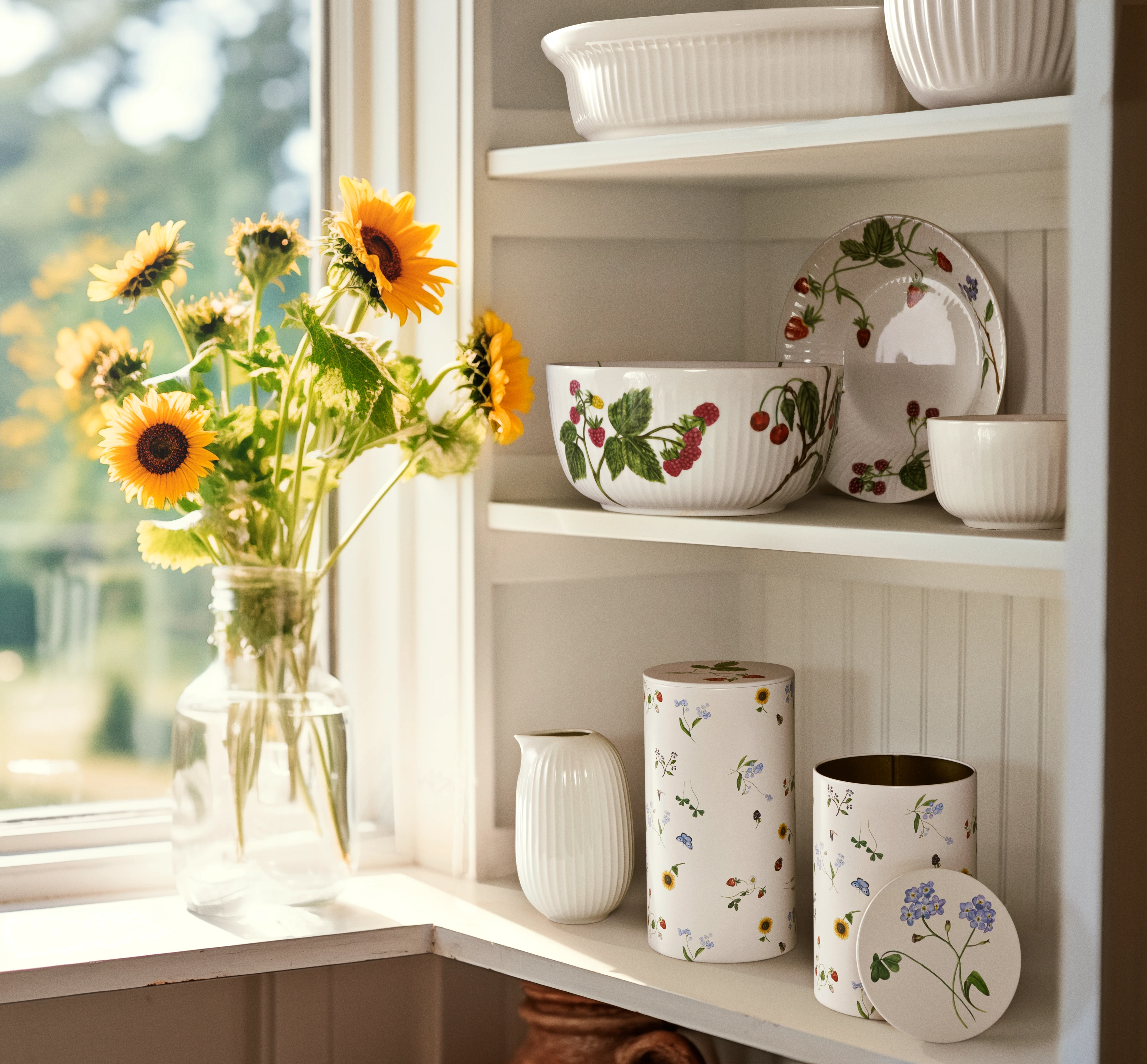 Kähler opbevaringsdåser på hvid hylde ved vindue med solsikker i vase og håndmalet porcelæn.