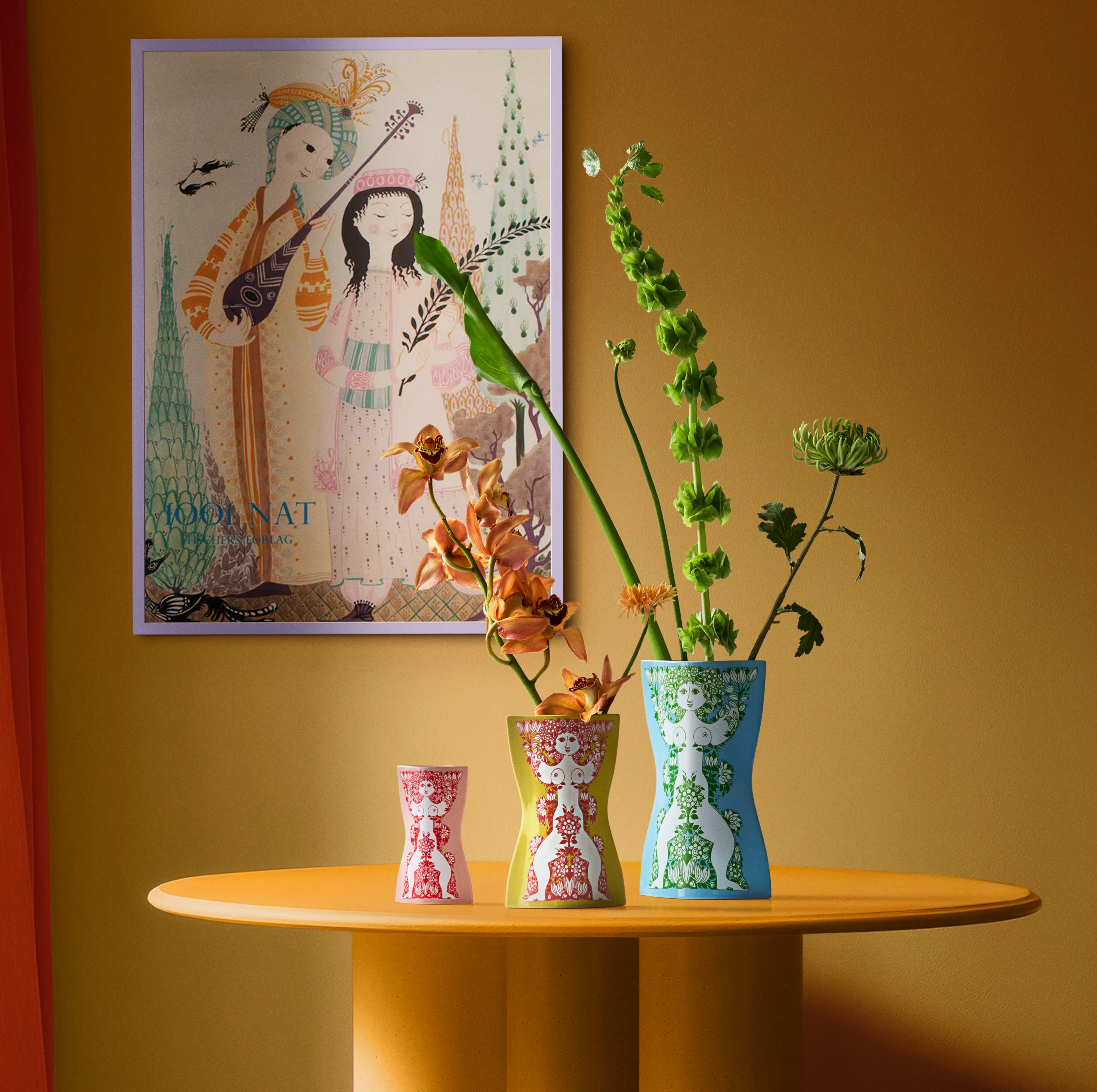 Bjørn Wiinblad Nina vaser med blomster på et rundt bord foran fargerik veggkunst.