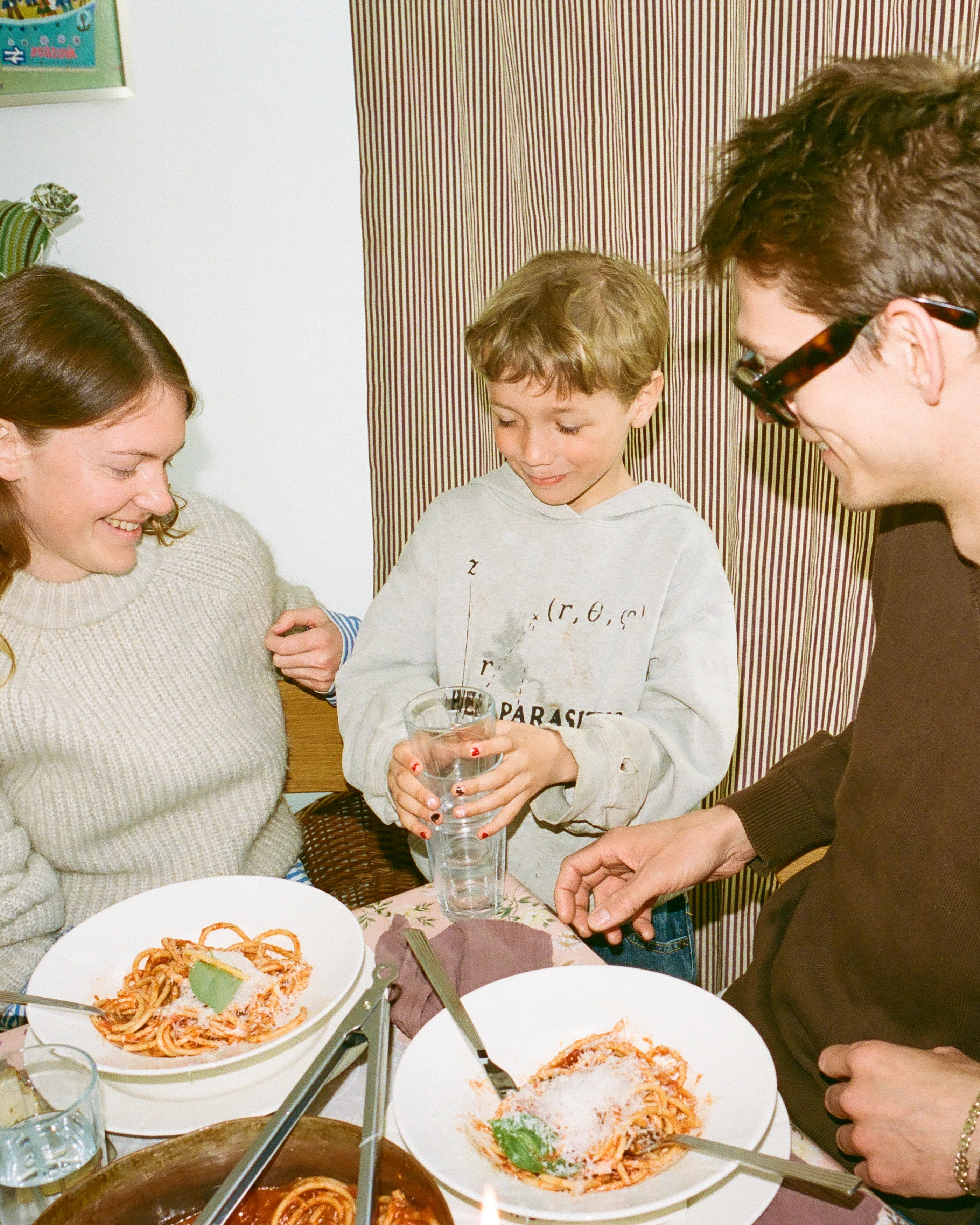 A warm family moment at the dinner table, with a child pouring water into a Rosendahl glass, surrounded by plates of pasta and smiling faces.
