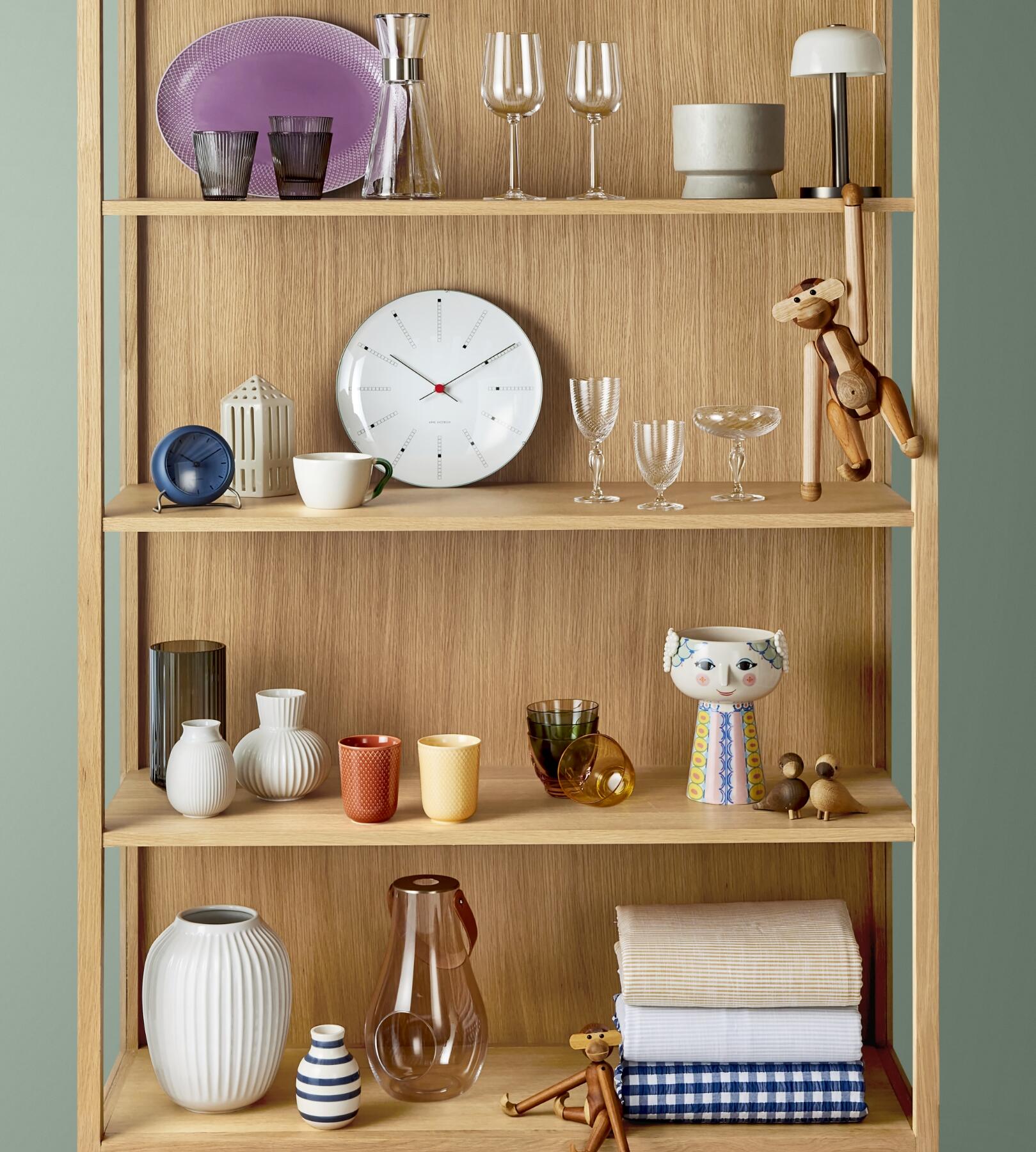 Shelf with a Bjørn Wiinblad vase, an Arne Jacobsen Clock, a Kay Bojesen Ape, wine glasses from Homegaard, a big white vase from Kähler, two colorful mugs from Lyngby and towels from Juna.